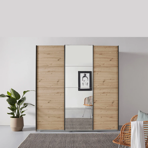 Pendle Village Furniture presents Rauch Forms 3 Door Sliding Wardrobe in Warm Oak with Metallic Grey Handles. Three internal compartments offer ample storage. Choose from two height options – 210cm or 229cm. Available in two width sizes: Small (203cm) and Large (271cm). Enjoy the convenience of Free Assembly - Middle Mirror Door Lifestyle Image